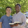 A boy wearing an orange and blue plaid shirt and holding an open book stands next to another boy wearing a blue shirt.  Photo credit: Kevin Hudson