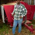 A man wearing a green and white plaid shirt, blue jeans, and a brown baseball cap stands in front of a bright red machine parked inside a metal building.  