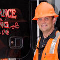 A man wearing a bright orange construction vest and hard hat stands in front of a logging machine.