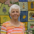 An elderly woman wearing an orange striped shirt stands in front of a large, multicolored, needlepoint county map of Mississippi.