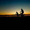 The silhouettes of two cyclists are seen in front of a blue and yellow sunrise that reflects on a lake below.