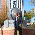 A man wearing a Jackson State University scarf and a navy suit jacket smiles in front of a sculpture on the JSU campus.