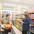 A woman wearing a Mississippi State University mask happily speaks with a woman pushing a grocery cart down an aisle of cereal in a grocery store.