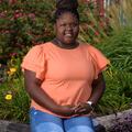 A smiling young girl wearing an orange shirt sits on a ledge in front of flowers.