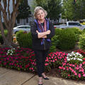 A woman, wearing all black clothes and a red, white, and blue scarf, smiling in front of flower bushes with her arms crossed.