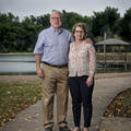A man with his arm around a smiling woman standing on a sidewalk in front of a lake.