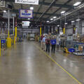A line of four people walking in a room full of machinery and boxes.