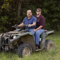 A man wearing a blue shirt and a woman wearing a maroon shirt sitting on a four-wheeler in tall green grass in front of dark green trees.