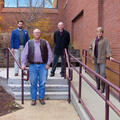 Three men and one woman standing, spaced out in front of a red brick building.