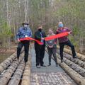 Four people stand behind a recently cut large red ribbon on a new bridge over a body of water.
