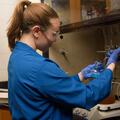 A young woman wearing safety goggles and a blue lab coat working in a lab.
