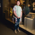 A smiling young girl wearing a floral blouse standing in front of a military museum exhibit.