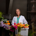 A woman wearing a white coat stands behind floral arrangements and a bucket full of flowers.
