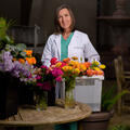 A woman wearing a white coat stands behind floral arrangements and a bucket full of flowers.