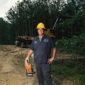 A man wearing a yellow hard hat and holding a chain saw stands in front of a trailer holding harvested timber.