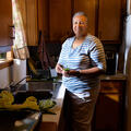 A woman wearing a white and blue striped shirt and holding fresh vegetables stands in front of a kitchen sink.