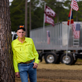 A man wearing a bright yellow button-up shirt leans against a large truck and rests one hand in his blue jean pocket.