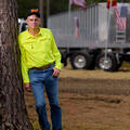 A man wearing a bright yellow button-up shirt leans against a large truck and rests one hand in his blue jean pocket.
