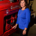 A smiling woman wearing a blue shirt stands next to and rests her arm on a red piece of machinery. 