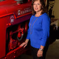 A smiling woman wearing a blue shirt stands next to and rests her arm on a red piece of machinery. 