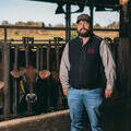A man gazes off to the left as the sun sets behind him and two cows look at him from the opposite side of the fence he is leaning on.