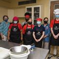 Five young adults and two women wearing black aprons and face masks smile for a group photo in a kitchen.