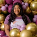 A smiling young woman wearing a shirt that reads “Beyond the Arch” stands next to several balloons and jars of party supplies.
