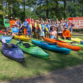 A large group photo of several young children and adults wearing kayaking gear and several kayaks of different colors in the grass in front of them. 