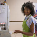 A smiling young girl wearing a green apron holding a measuring cup full of flour.
