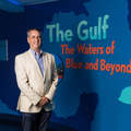 A man wearing dress clothes stands in front of a blue wall and smiles with his hands clasped in front of his body.