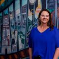 A smiling woman in a blue shirt stands in front of a wall mural.