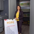 A professionally dressed woman standing in front of many rows of food cans and beside a sign that lists “HOPE.”