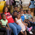 A woman, wearing a red T-shirt listing Rosemary’s Daycare on it, smiles while sitting on a bench with many small children sitting beside her, also smiling.