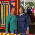 Two Black women smiling and standing in front of a hanging multicolored quilt.