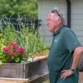 A man, standing beside a wooden table filled with plants, looking into the distance.