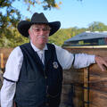 A person with a gray mustache and wearing a black cowboy hat, white polo, and black vest with a sheriff’s star on his chest.