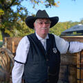 A person with a gray mustache and wearing a black cowboy hat, white polo, and black vest with a sheriff’s star on his chest.