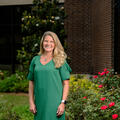A smiling person wearing green and standing outside in front of roses.