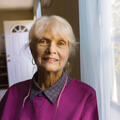 An smiling elderly woman stands next to a window. 