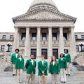 Seven teenagers wearing green blazers and tan slacks form a U standing on steps in front of six gray columns.