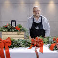 A man, smiling, standing in front of a candle centerpiece and behind a table with bows, evergreen door swag and evergreen garland.