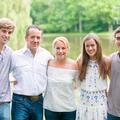 Family photo of 2 parents and 3 young adults standing with arms around each other in front of a small body of water.