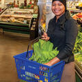 A woman in a grocery store holding a blue basket filled with produce like leafy green lettuce.