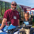 A man stands next to an ATV with a young boy sitting on the seat.