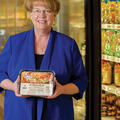 A smiling woman wearing a blue shirt stands in front of a frozen food aisle in a grocery store.