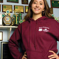 A girl wearing a maroon sweatshirt stands in front of a trophy display with her hands on her hips.