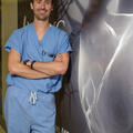 A man in blue scrubs with crossed arms leans against a wall.