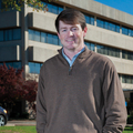 A man with hands in his pockets stands smiling in front of a large concrete building.