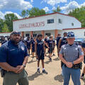 Two officers stand in front of a group of young men spaced apart for workouts in a parking lot.