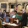 A young woman wearing a dark green blazer stands behind a podium while several people stand looking at her. 